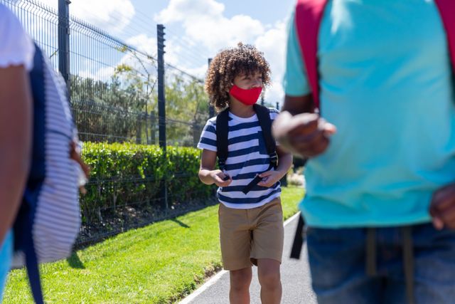 African American boy wearing a face mask and backpack walking on a footpath. Ideal for use in educational materials, health and safety campaigns, and articles about school life during the coronavirus pandemic.