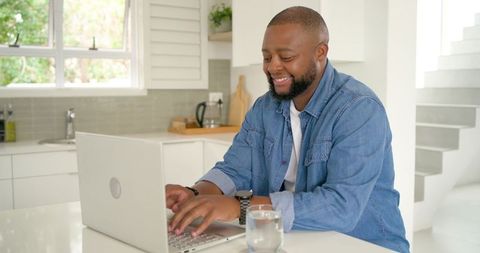African american man in thirties smiling while working remotely on laptop at bright kitchen island
