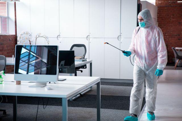 Caucasian male wearing protective coverall, mask, and gloves disinfecting modern office space. Ideal for illustrating workplace hygiene, COVID-19 safety measures, professional cleaning services, and health precautions in business environments.