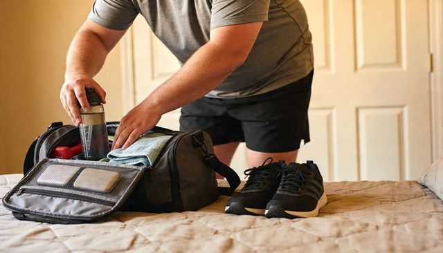 Man packing gym duffel bag on bed with shaker bottle, towel and sneakers