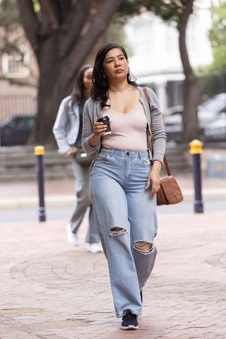 Young woman walking holding coffee cup and crossbody bag wearing ripped jeans on city sidewalk