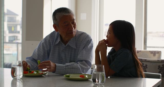 Older man and young girl enjoying healthy snacks with plates of vegetables at kitchen counter. This image can be used to highlight family bonding, healthy lifestyles, and intergenerational relationships. Suitable for articles or advertisements promoting family time, nutrition, and balanced eating habits.
