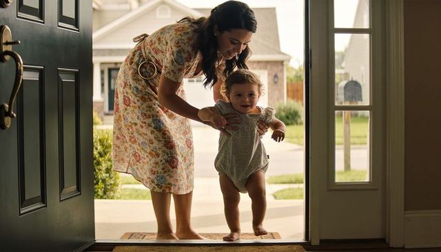 Mother guiding toddler stepping over threshold on sunny porch — family home moment