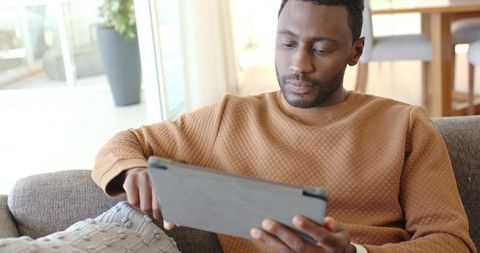 African American man relaxing on sofa using tablet in sunlit modern living room