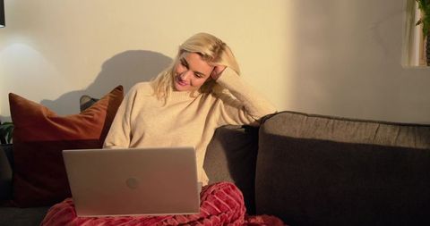 Sunlit blonde woman relaxing on sofa with laptop and cozy blanket