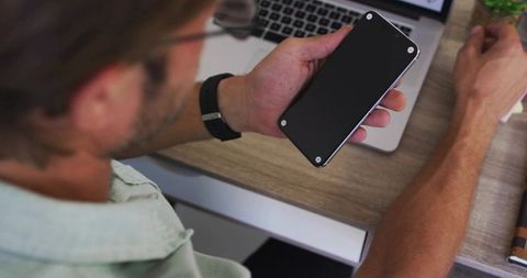 Man holding smartphone and scrolling at wooden desk with laptop, planner and wristwatch