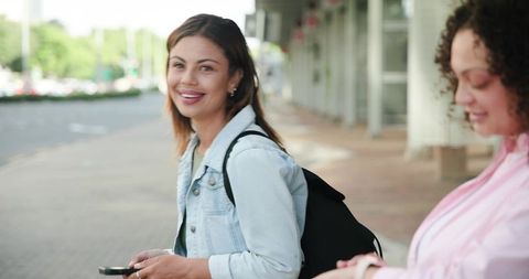 African American women smiling on urban sidewalk holding smartphone and backpack, daylight