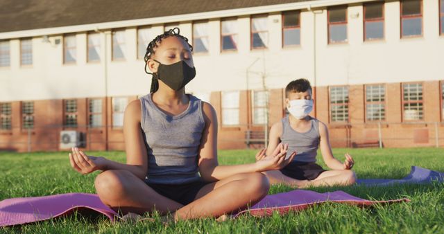 Two children in face masks meditate on yoga mats on school grounds. This can illustrate articles or marketing on mindfulness, health practices during the pandemic, exercise for kids, or educational activities promoting wellness.