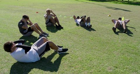 Outdoor fitness training: diverse male teammates doing sit-ups on grass with cones