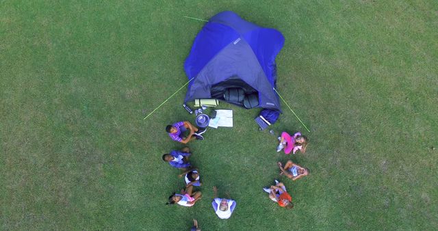 Aerial view of a family camping scene with a tent set up on grass. Eight people, including children, are sitting in a circle around the tent, engaging in conversation or planning activities. This shot is perfect for advertising camping gear, vacations, outdoor activities, or portraying family bonding in nature.