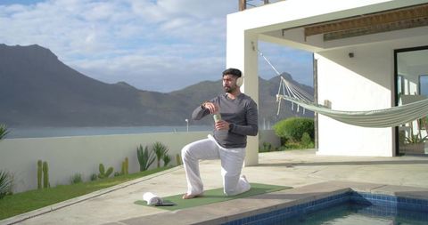 Indian man practicing yoga on poolside patio with headphones, water bottle, hammock view