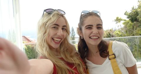Young women taking cheerful selfie on sunny balcony with sunglasses and city view