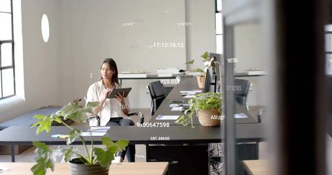 Asian woman using tablet while working at minimalist sunlit office desk with plants