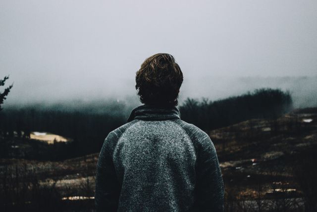Man dressed in dark clothing standing in foggy, mountainous wilderness. Visible back view and overcast atmosphere suggest solitude and contemplation. Ideal for concepts of reflection, adventure, exploring nature, and solitary moments in misty outdoors.
