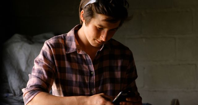 Depicts a young farmer in a plausible environment focusing on a smartphone. Suitable for illustrating the blending of modern technology with traditional farming practices, rural connectivity, or agricultural innovation. Can be used in articles, advertisements, or educational materials highlighting technology in agriculture or rural development.