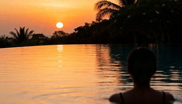 Woman gazing across infinity pool at tropical sunset with warm sun reflection