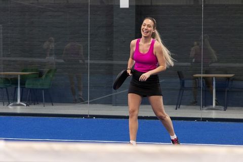 Smiling woman playing pickleball on indoor blue court in magenta top and black skirt