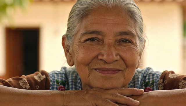 Senior hispanic woman leaning on porch railing smiling warmly wearing embroidered blouse