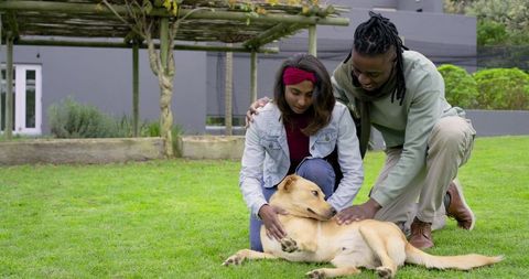 African american man and indian woman kneeling on lawn petting tan dog in modern backyard