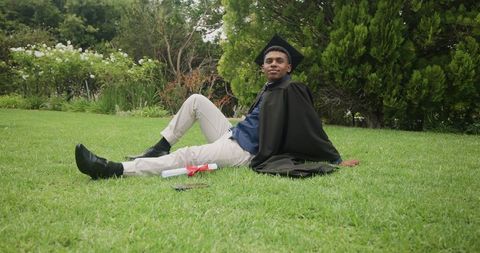 Relaxed african american graduate reclining on grass wearing cap and gown holding diploma