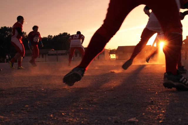 American football players engaging in a high-energy training session on a dusty field while the sun sets in the background. This scene captures the intensity, teamwork, and dedication of athletes. Ideal for use in sports-related promotions, teamwork and perseverance advertisements, or illustrating athletic training environments.