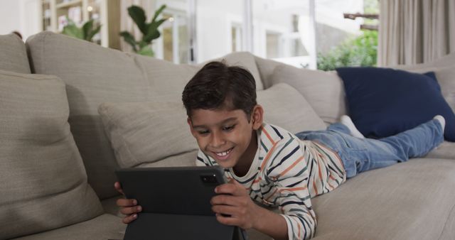 Young boy laying on couch and using tablet device, smiling while engaged in online activities. Ideal for content related to children's technology use, digital learning, remote education, online entertainment, and comfortable home environments.