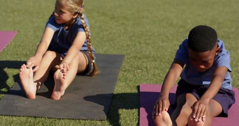 Diverse children stretching and reaching toes on yoga mats outdoors on sunny grass