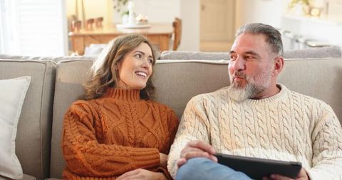 Middle-aged couple relaxing on sofa in cozy cable-knit sweaters using tablet