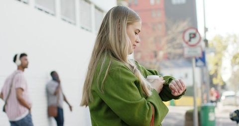 Young woman in green coat checking wristwatch while holding coffee on urban sidewalk