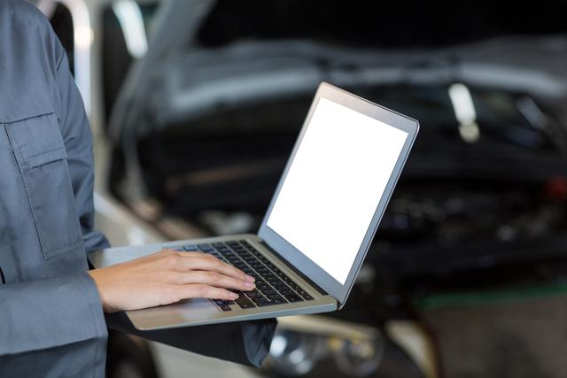 Female mechanic using laptop in repair garage, ideal for illustrating modern automotive repair, technology in car maintenance, and professional mechanics. Useful for articles, blogs, and advertisements related to automotive services, repair shops, and technical training.