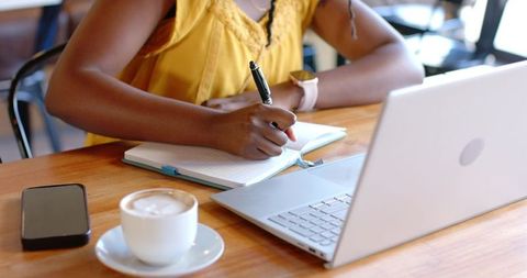 African woman writing in notebook at cafe with laptop and coffee, sunlit workspace