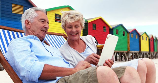 Senior Couple Relaxing on Beach with Tablet in Front of Colorful Beach Huts - Download Free Stock Images Pikwizard.com