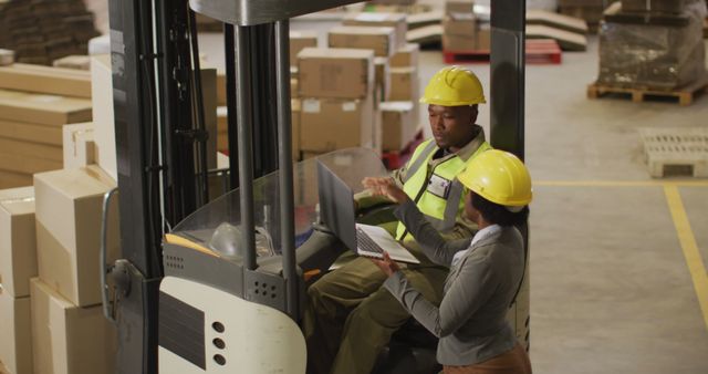 Two warehouse workers using a laptop for logistics management in a warehouse. Both are wearing safety helmets and safety vests, surrounded by boxes and parcels. This can be used to illustrate workplace collaboration, logistics planning, and warehouse operations.