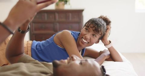 Smiling african american woman relaxing with partner on bed sharing phone and laughter