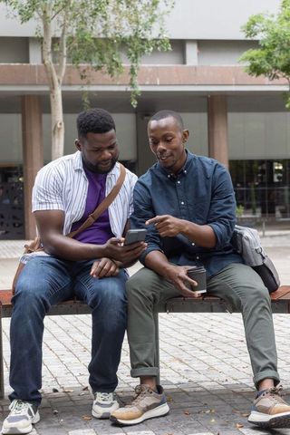 Two african men sitting on bench sharing smartphone and coffee in urban courtyard