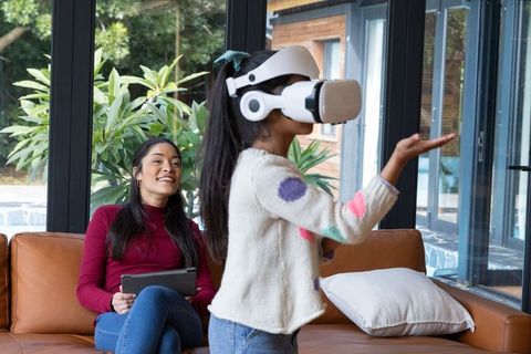 Asian mother watching daughter playing with white VR headset in bright modern living room