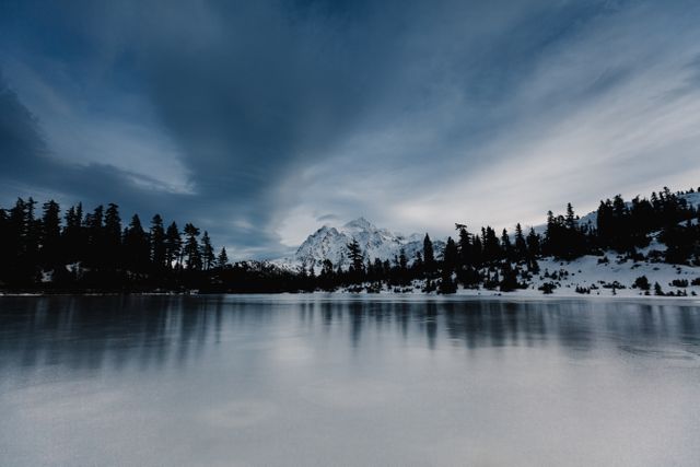 Winter landscape featuring an icy lake surrounded by a snow-covered forest and majestic mountains under a cloudy sky. Ideal for use in nature photography, winter travel promotions, holiday greeting cards, outdoor adventure blogs, and winter-themed home decor.