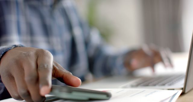 Image shows a close-up of a person's hand using a smartphone while working at a desk. A laptop and documents are visible. Ideal for themes related to business, technology, multitasking, productivity, and remote work.