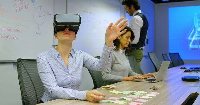 A woman is engaging with a virtual reality headset in a modern office, suggesting an interactive and innovative business meeting. Colleagues are busy working with laptops and notes. This image is ideal for illustrating concepts of technology integration, future workspaces, business innovation, and team brainstorming sessions.