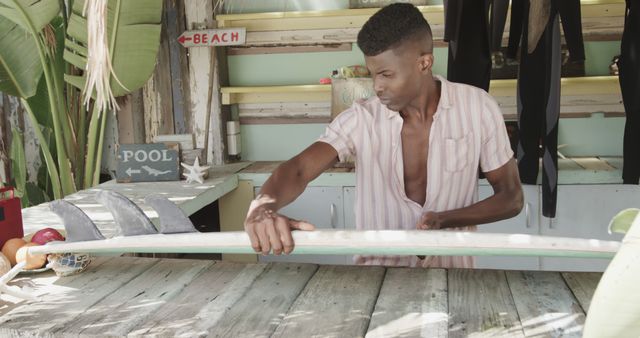 Focused african american man in surf shop on beach preparing surfboard. Lifestyle, business, vacation, summer and leisure, unaltered.