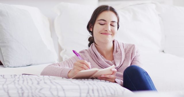 A young woman is happily journaling while sitting on the floor in a cozy room. The setting exudes relaxation and comfort, making it suitable for themes around leisure, self-care, mindfulness, and emotional well-being. This image could be used for articles or blog posts about journaling, mental health, relaxation techniques, or lifestyle content.