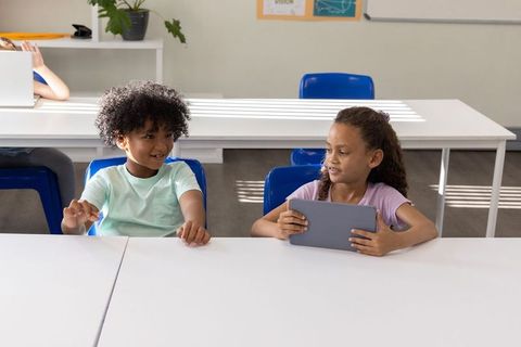 Diverse schoolchildren using tablet and chatting at classroom table