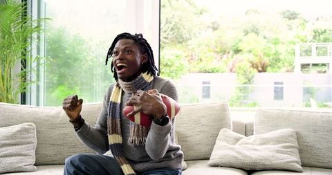Excited African American Man Cheering While Holding Football on Modern Living Room Sofa