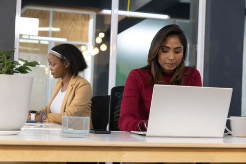 Indian professional and coworker working in open-plan office with laptop and paperwork