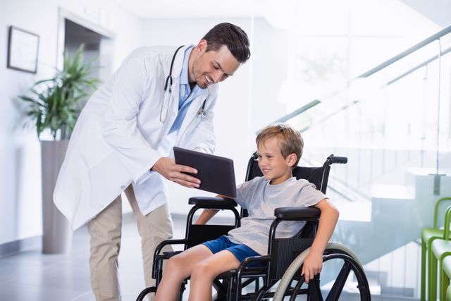 Doctor showing digital tablet to boy in wheelchair in hospital corridor. Ideal for use in healthcare, medical technology, pediatric care, and patient interaction contexts. Can be used in articles, brochures, and websites promoting medical services, pediatric care, and healthcare technology.