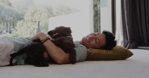Smiling man lying on carpet hugging brown dog while resting on mustard pillow