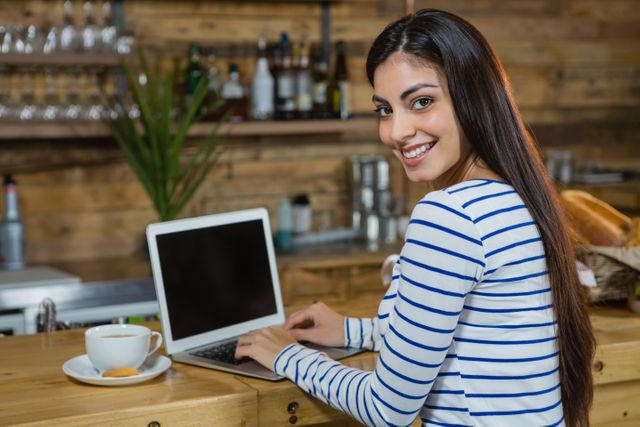 Young woman smiling while using laptop at cafe counter. Ideal for illustrating remote work, modern lifestyle, casual work environments, and technology use in everyday life. Suitable for blogs, websites, and advertisements related to coffee shops, freelancing, and digital nomadism.
