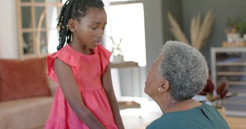 African american grandmother listening to granddaughter in sunlit living room wearing coral dress