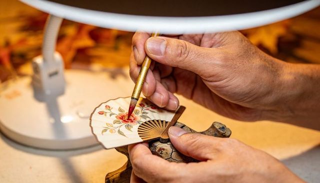 Artist hand-painting miniature folding fan with detailed red flower on workbench under lamp