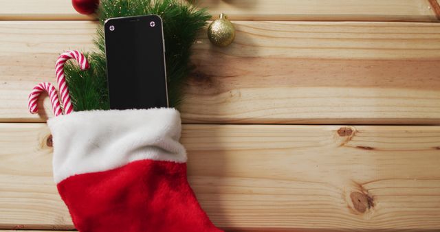 Smartphone with two candy canes tucked in red Christmas stocking on wooden table with golden ornaments. Perfect for festive-themed advertisements, holiday gifting promotions, or Christmas decorations marketing.